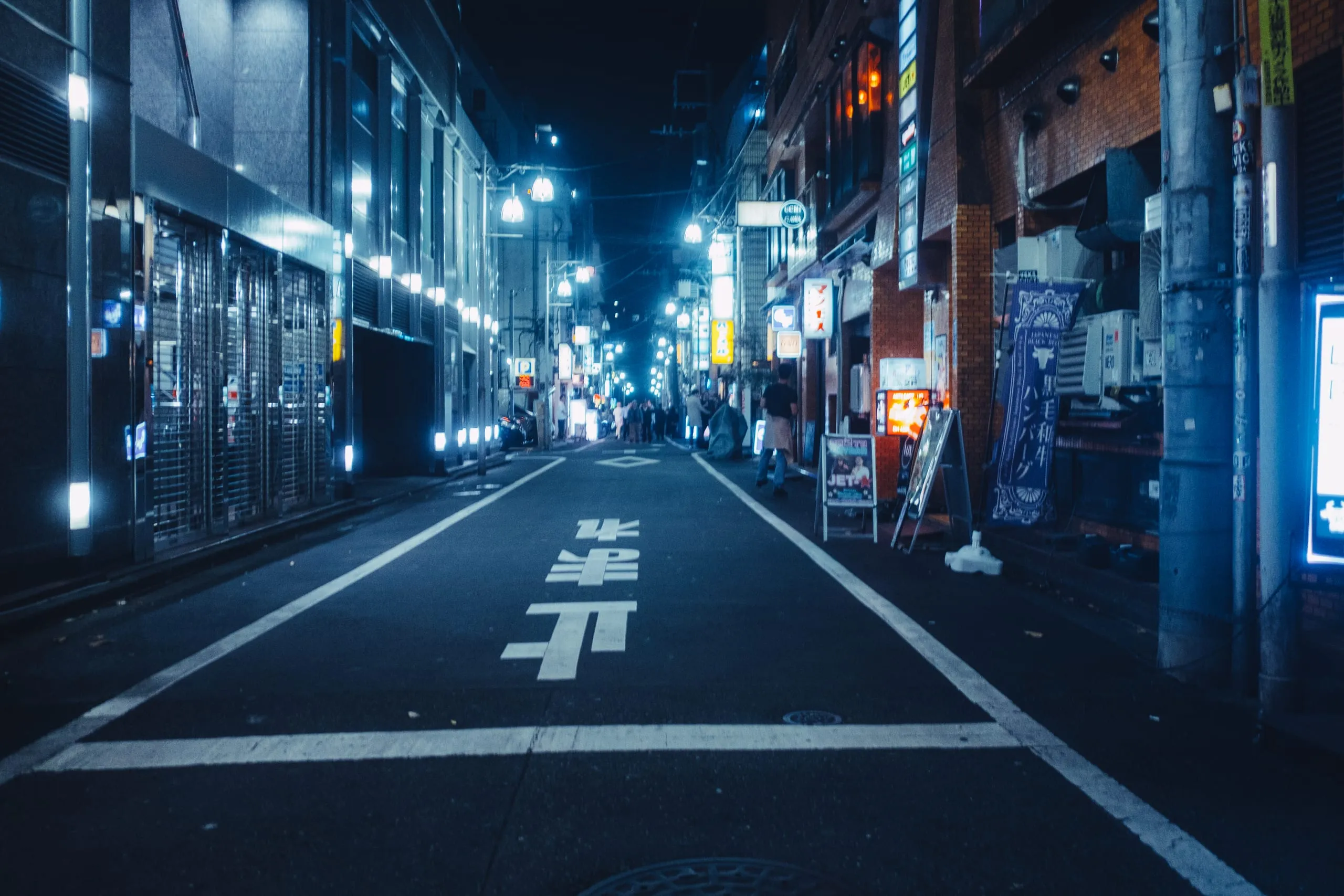 An empty backstreet lit blue by lamps and signs at night