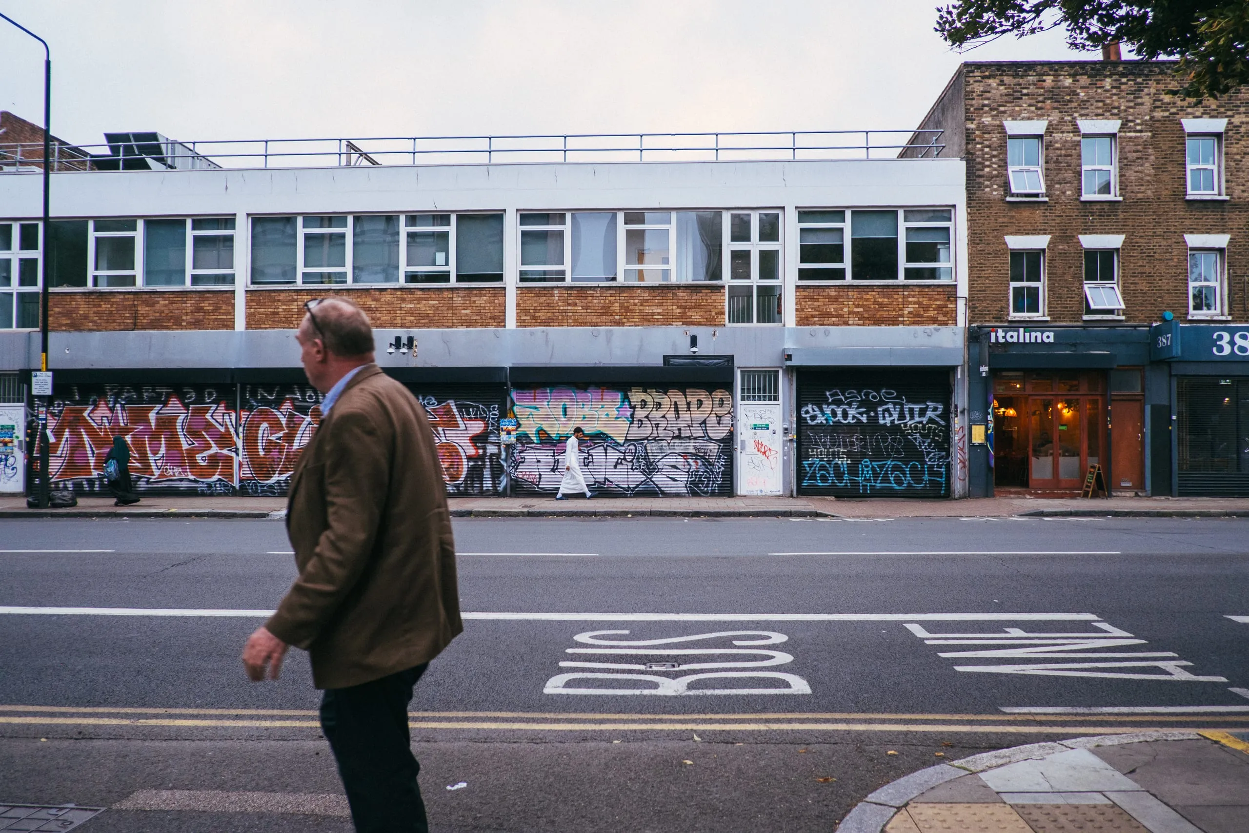 A man crosses the road past a graffiti-covered shopfront and bus lane, Bethnal Green
