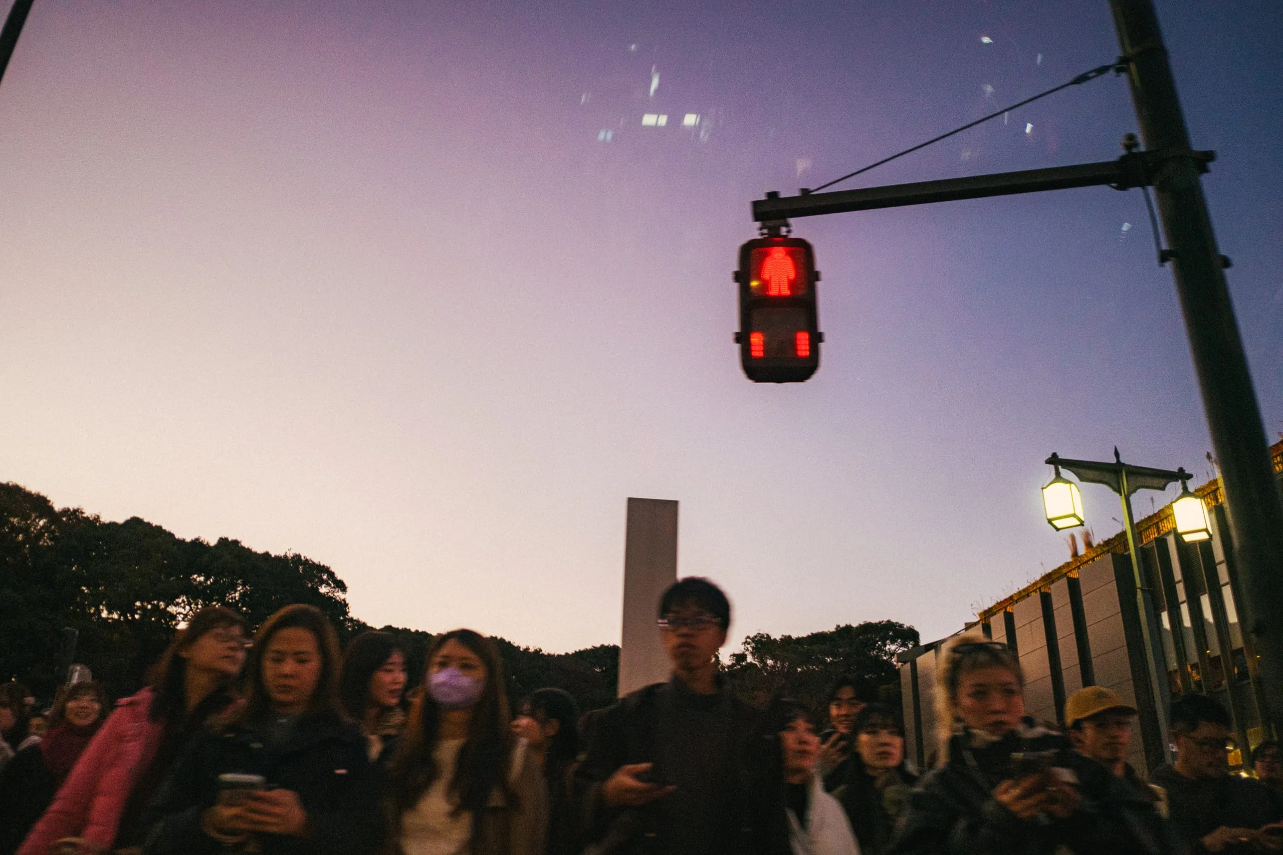 A crowd waits at a red pedestrian signal at dusk