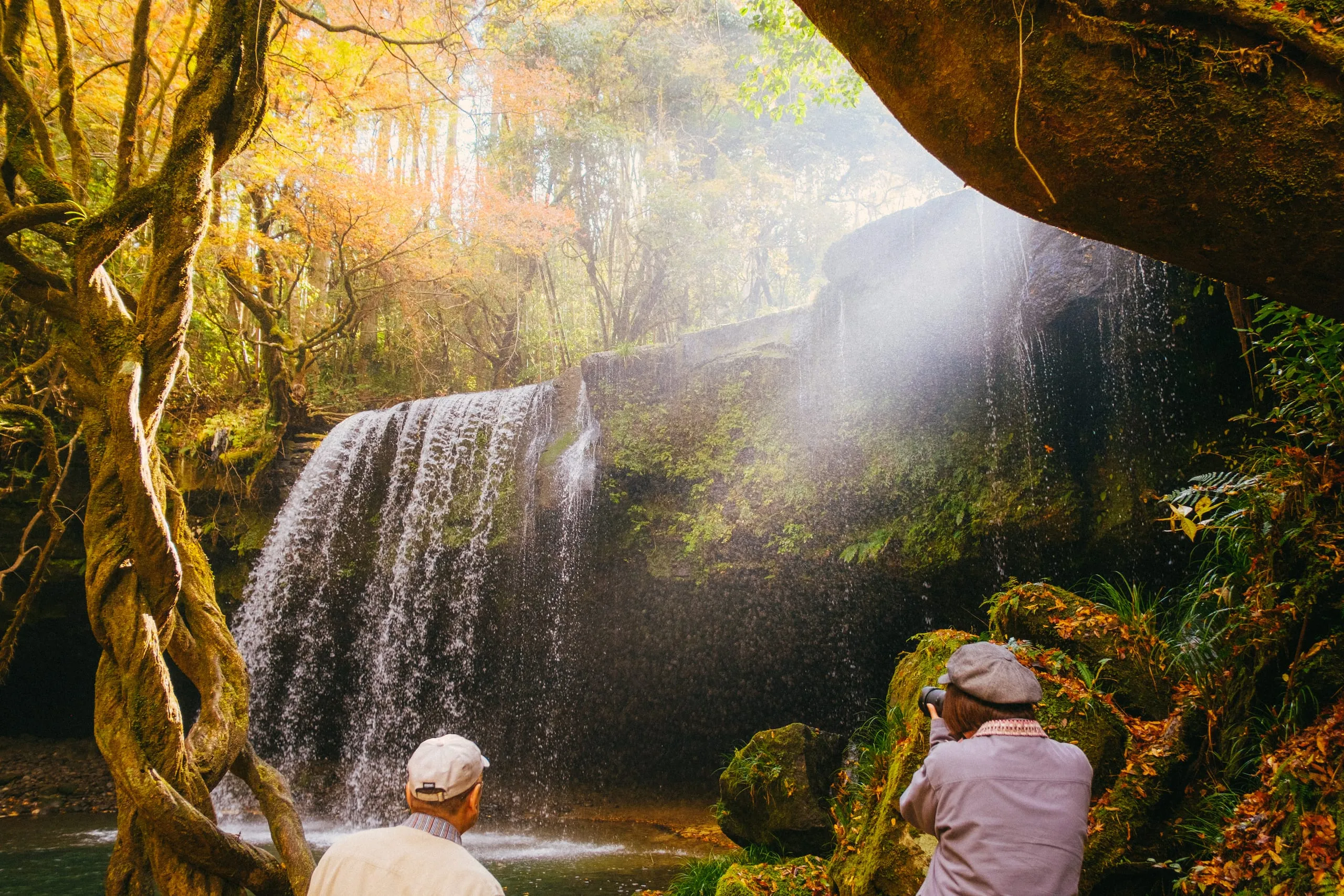 An elderly couple photograph a waterfall in autumn woodland