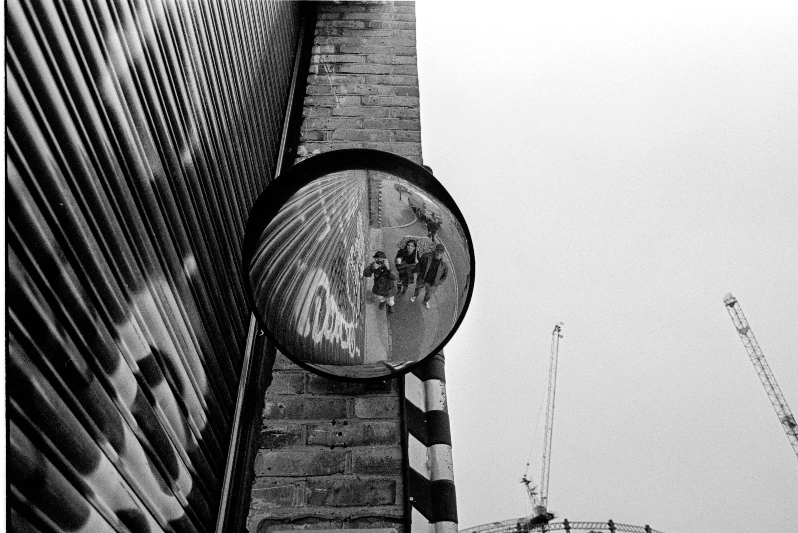 People reflected in a convex traffic mirror beside a shuttered building, with construction cranes behind