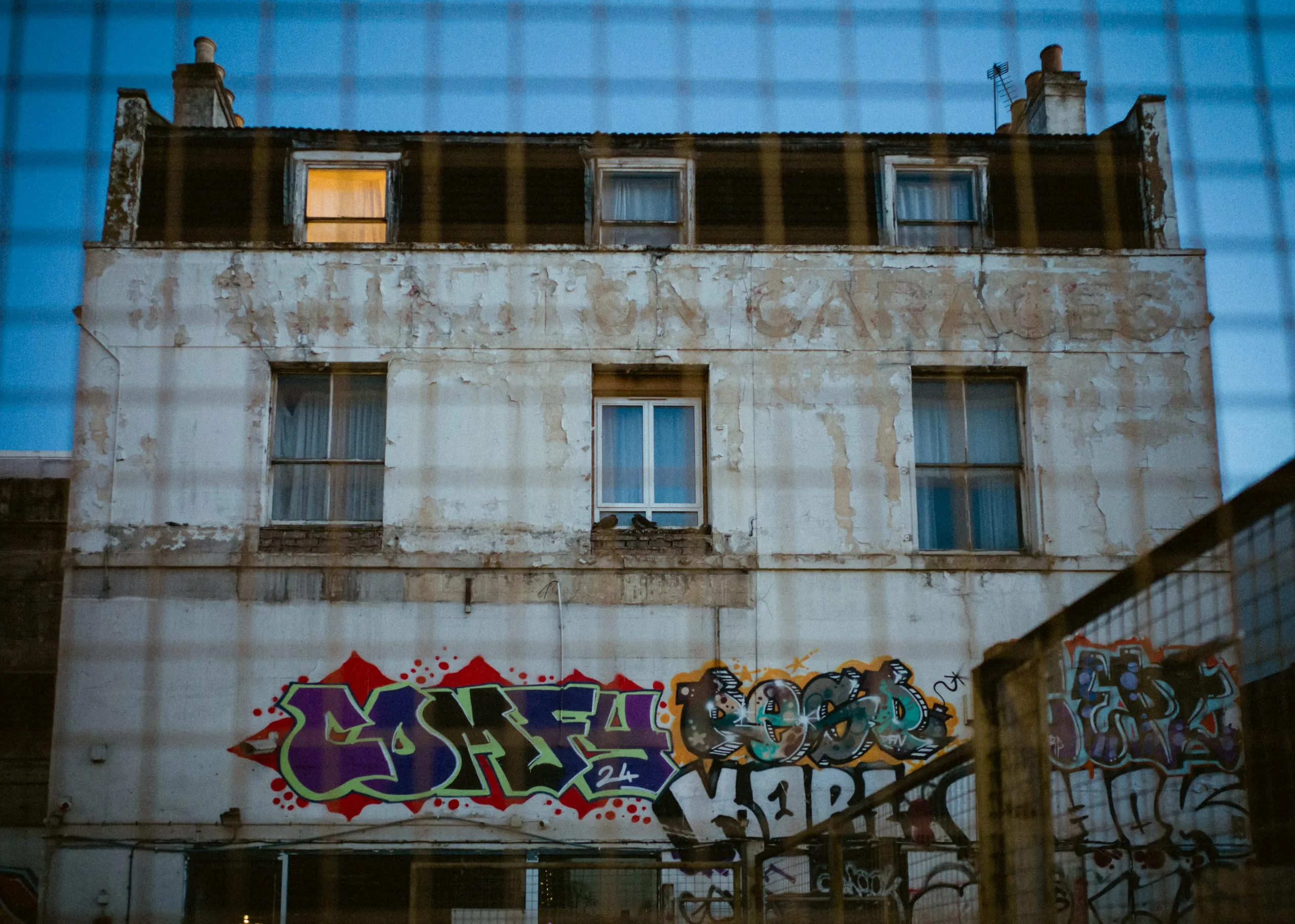 Graffiti on the wall of a derelict garage building at dusk, seen through a fence
