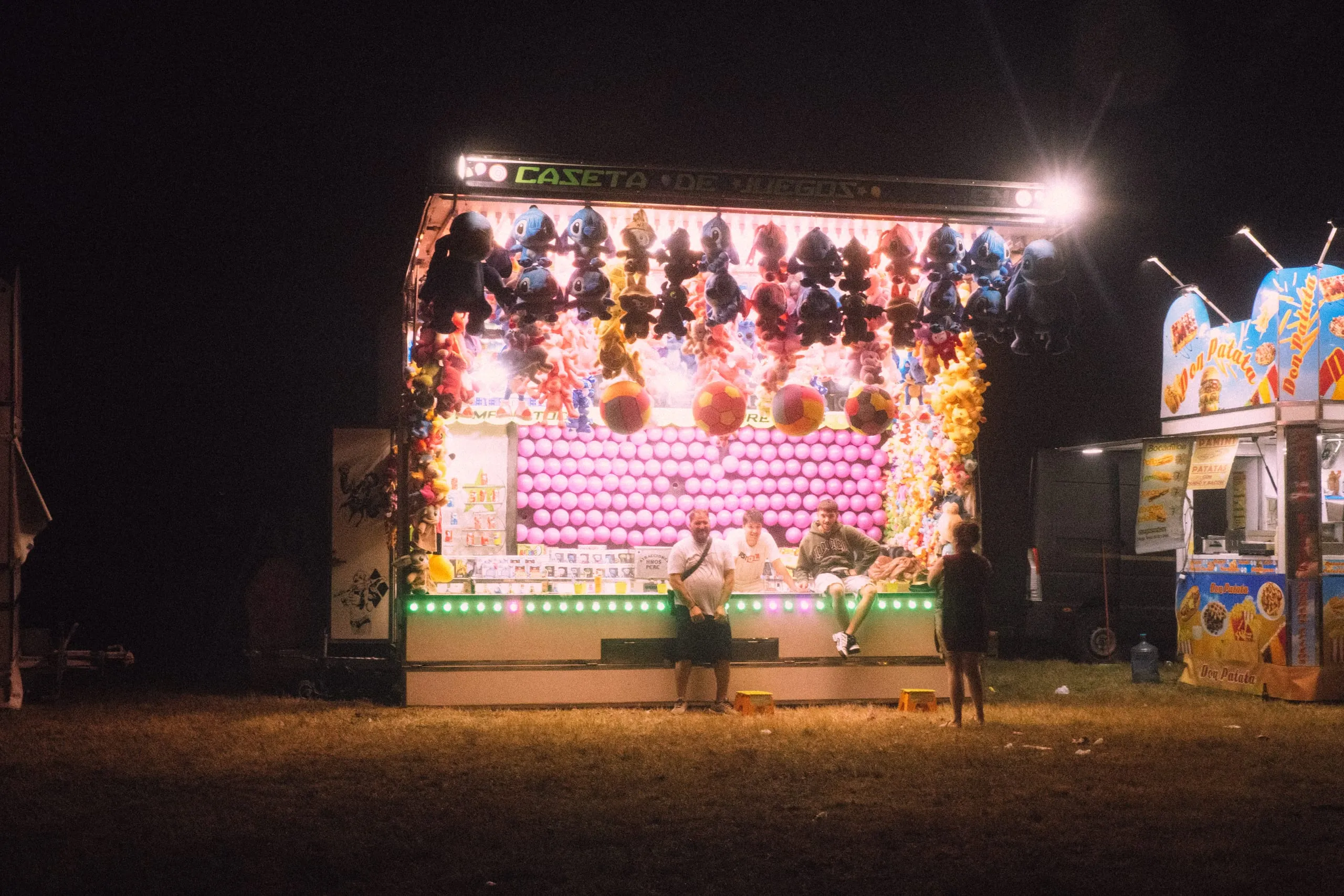 People sit at a lit fairground game stall at night en las Fiestas de Lugo, Llanera