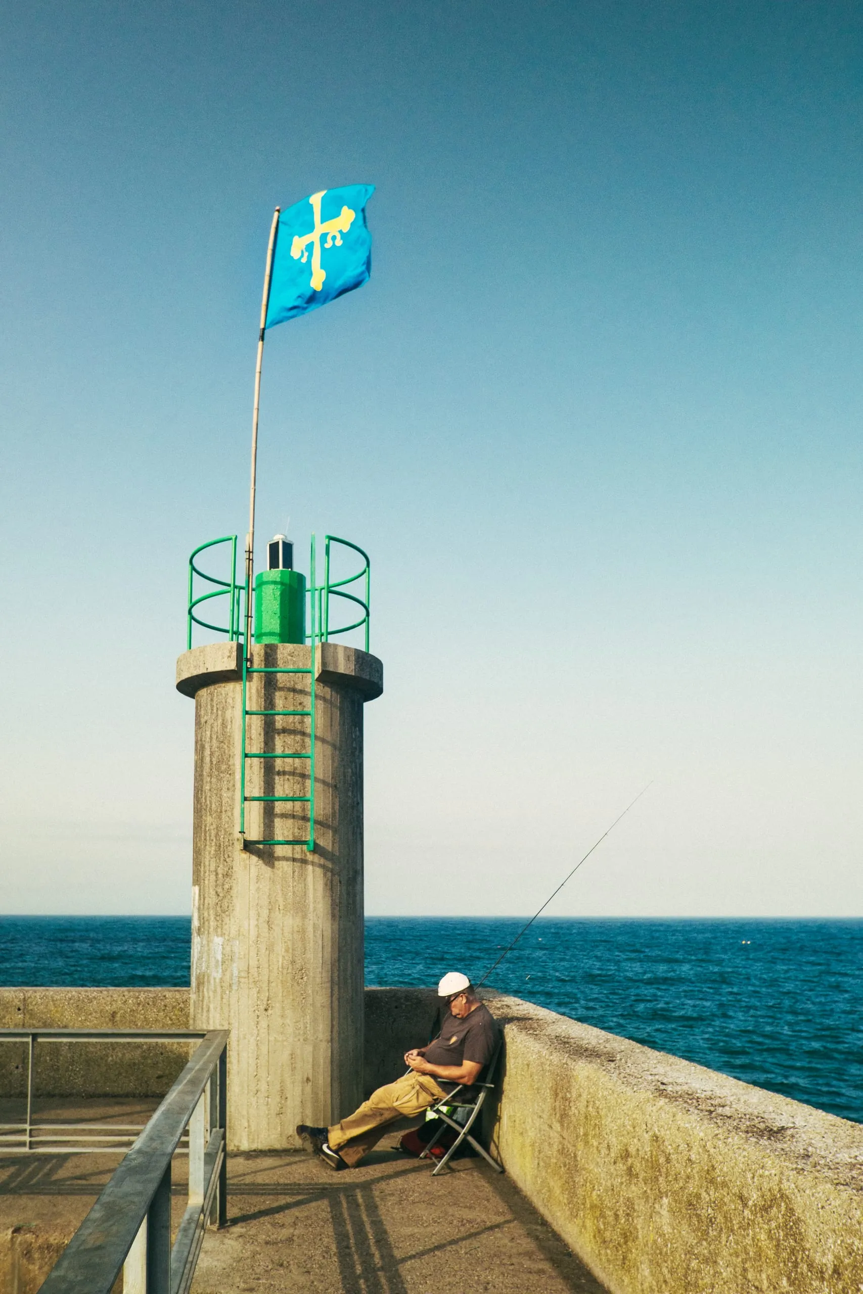 A man fishes from a breakwater beneath an Asturian flag, Tazones