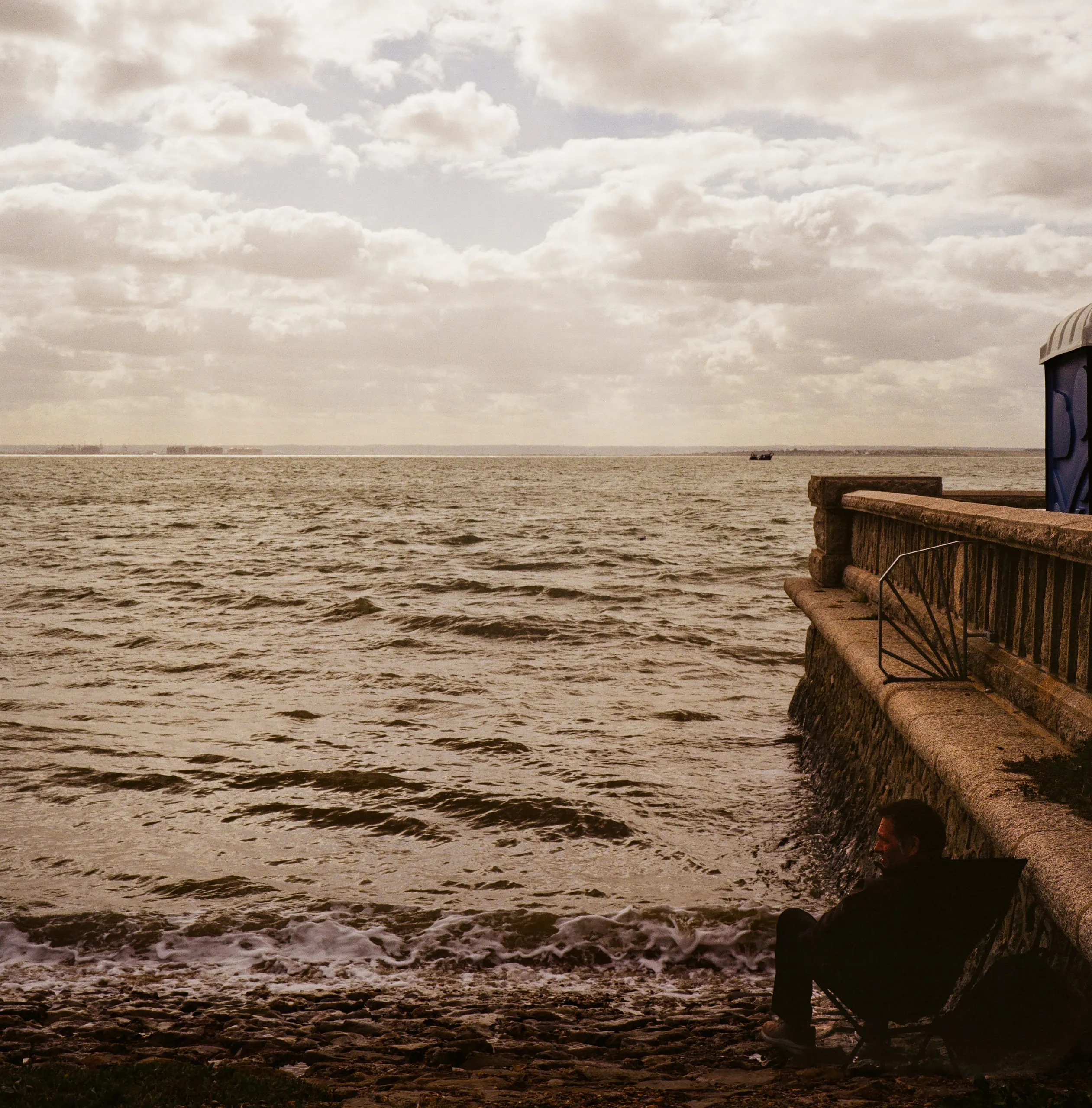 A man sits against a sea wall beside the choppy water on a windy day