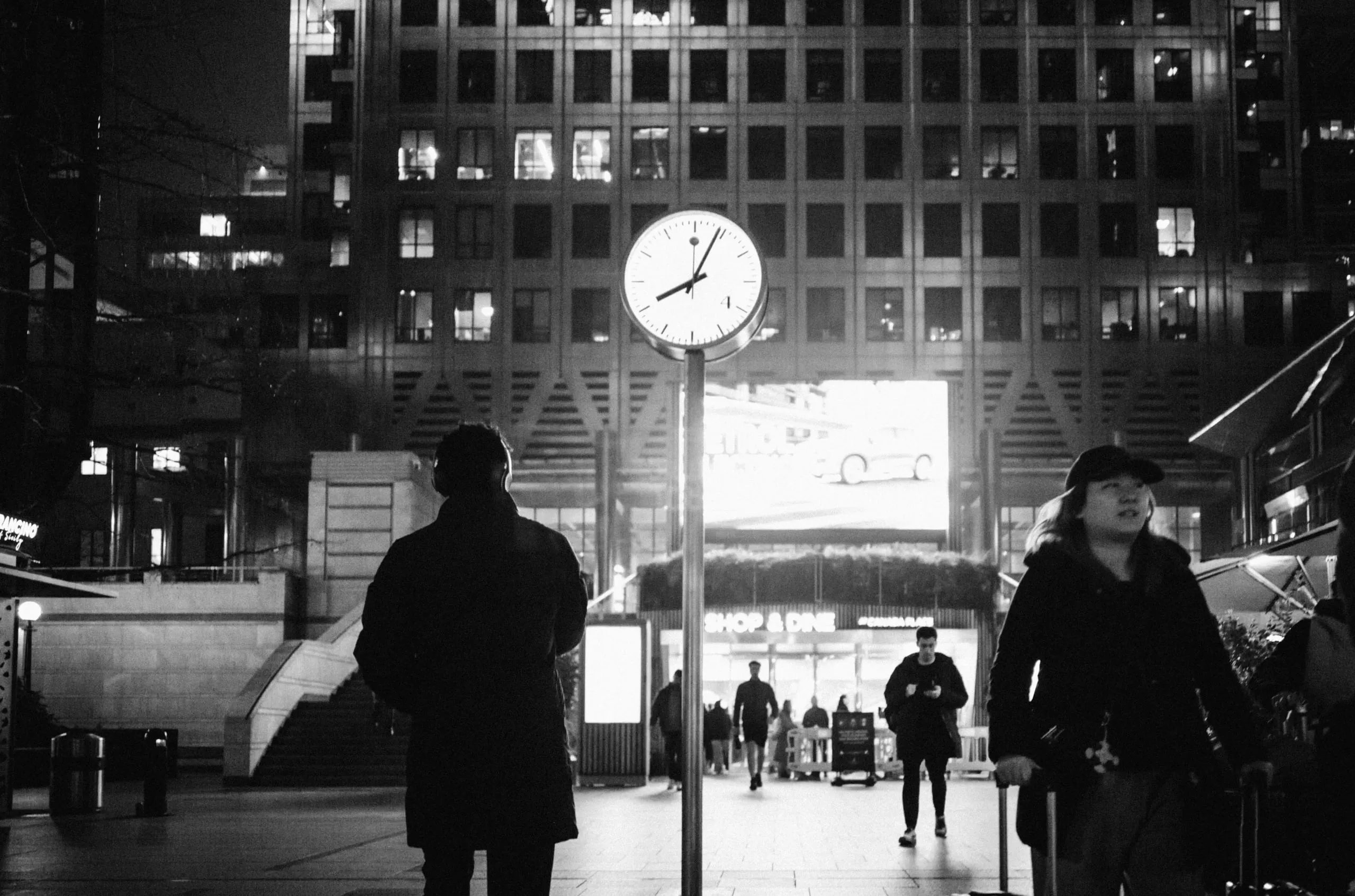 People pass a clock on a pole in a lit plaza at night, black and white, Canary Wharf