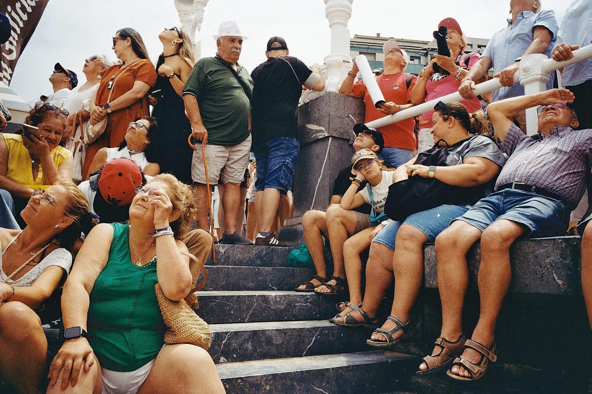 A man in a green polo shirt stands at the top of a flight of stone steps, surrounded by spectators sitting and standing, most looking up at the sky
