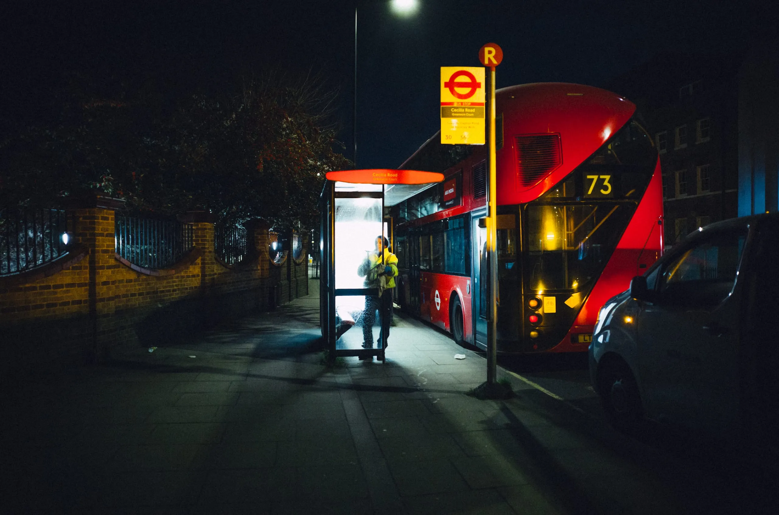 A worker stands lit inside a bus shelter beside a route 73 bus at night, London