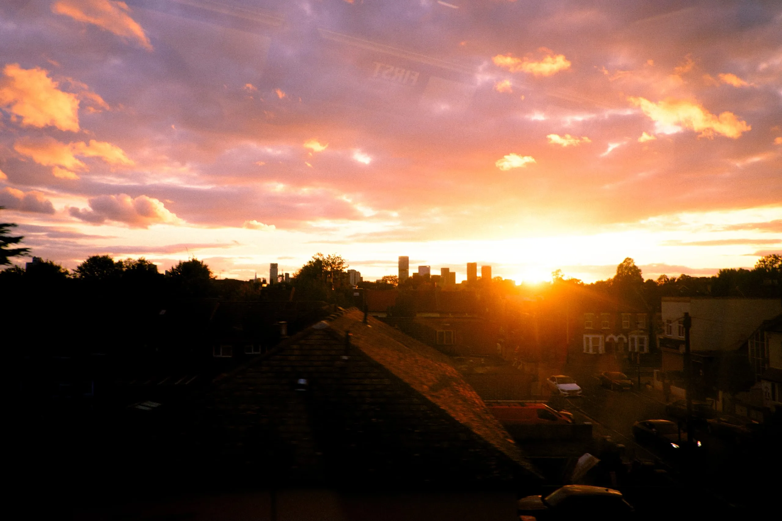 The sun sets behind the rooftops and distant tower blocks of a city