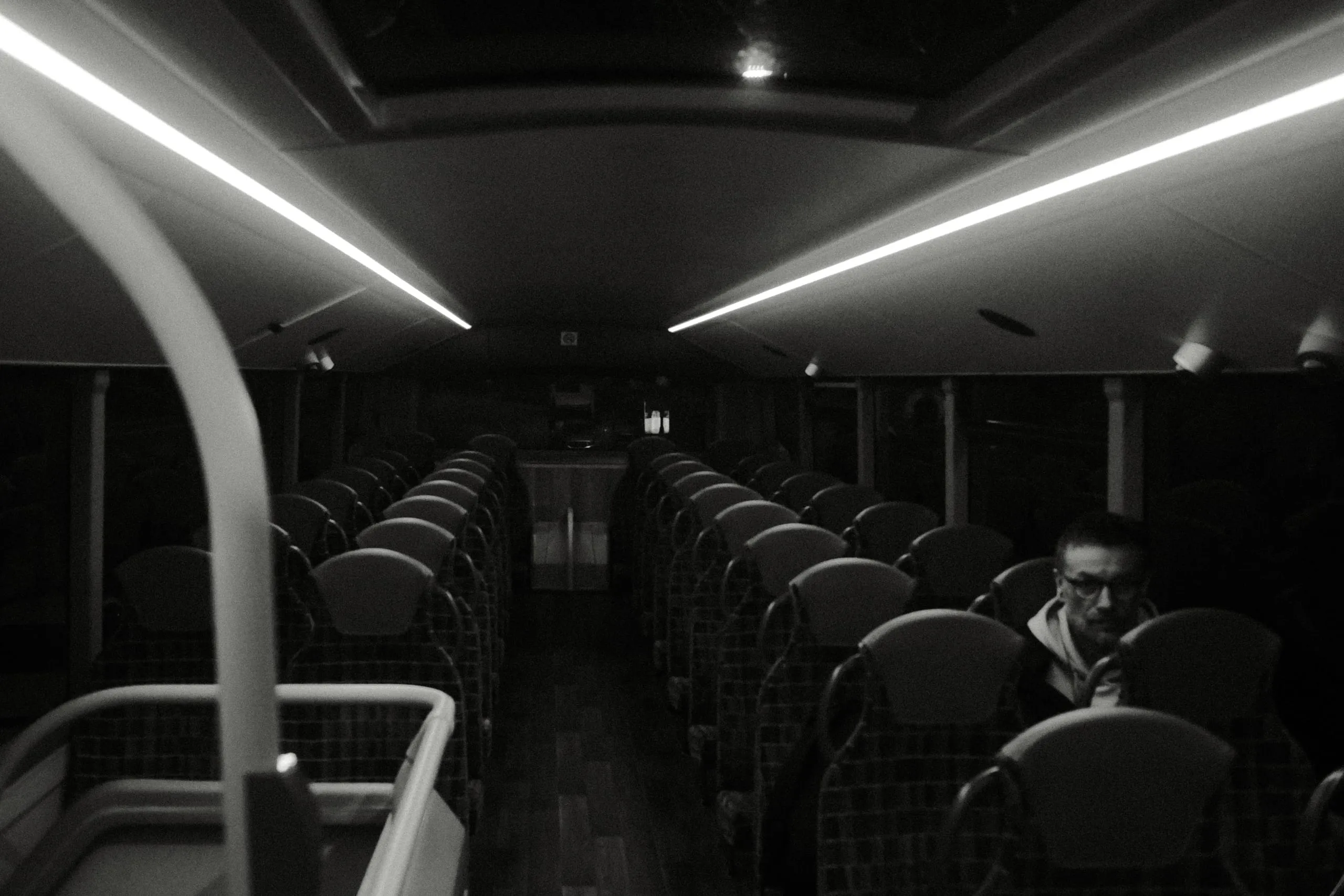 A lone passenger sits among empty seats on the upper deck of a bus at night
