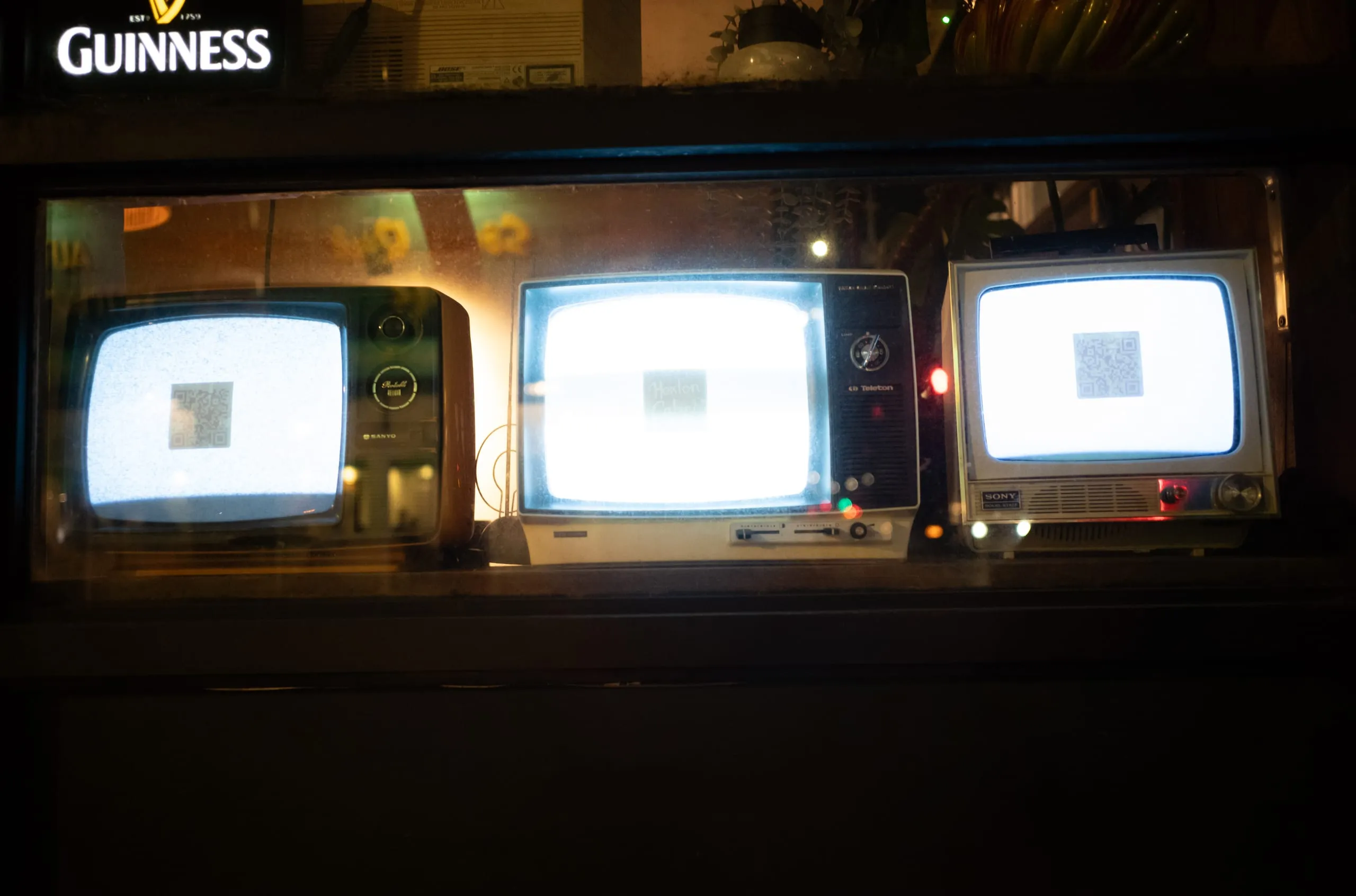 Three vintage televisions displaying QR codes in a shop window below a Guinness sign