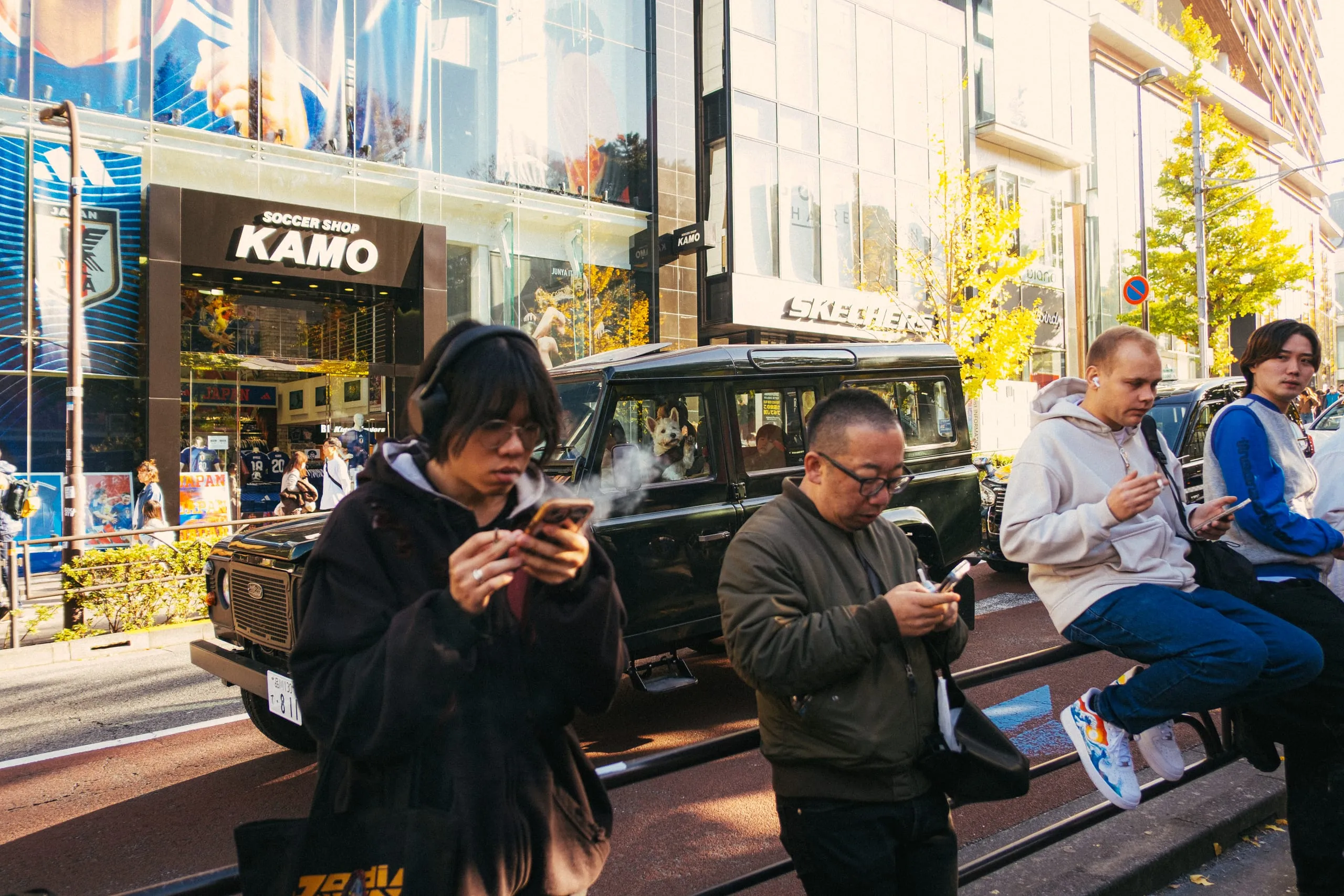 Smokers check their phones while a west highland terrier is driven past Harajuku station