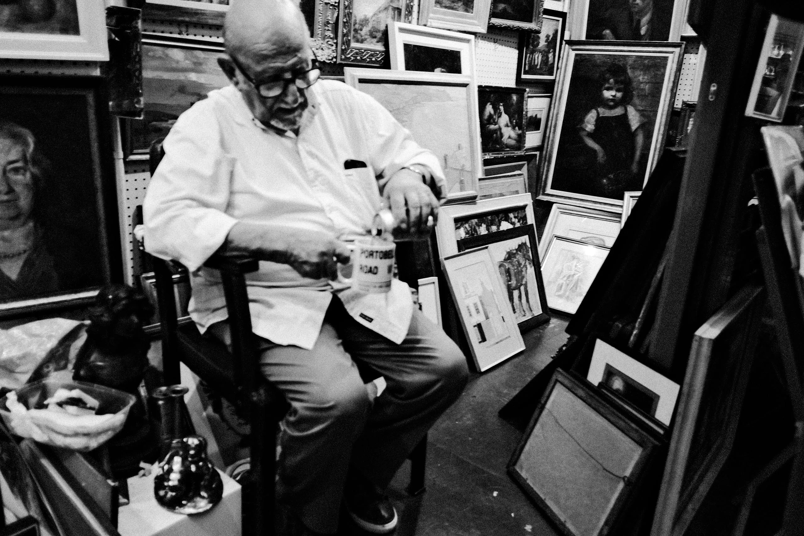 An older man sits among framed paintings and prints at a stall, Portobello Road