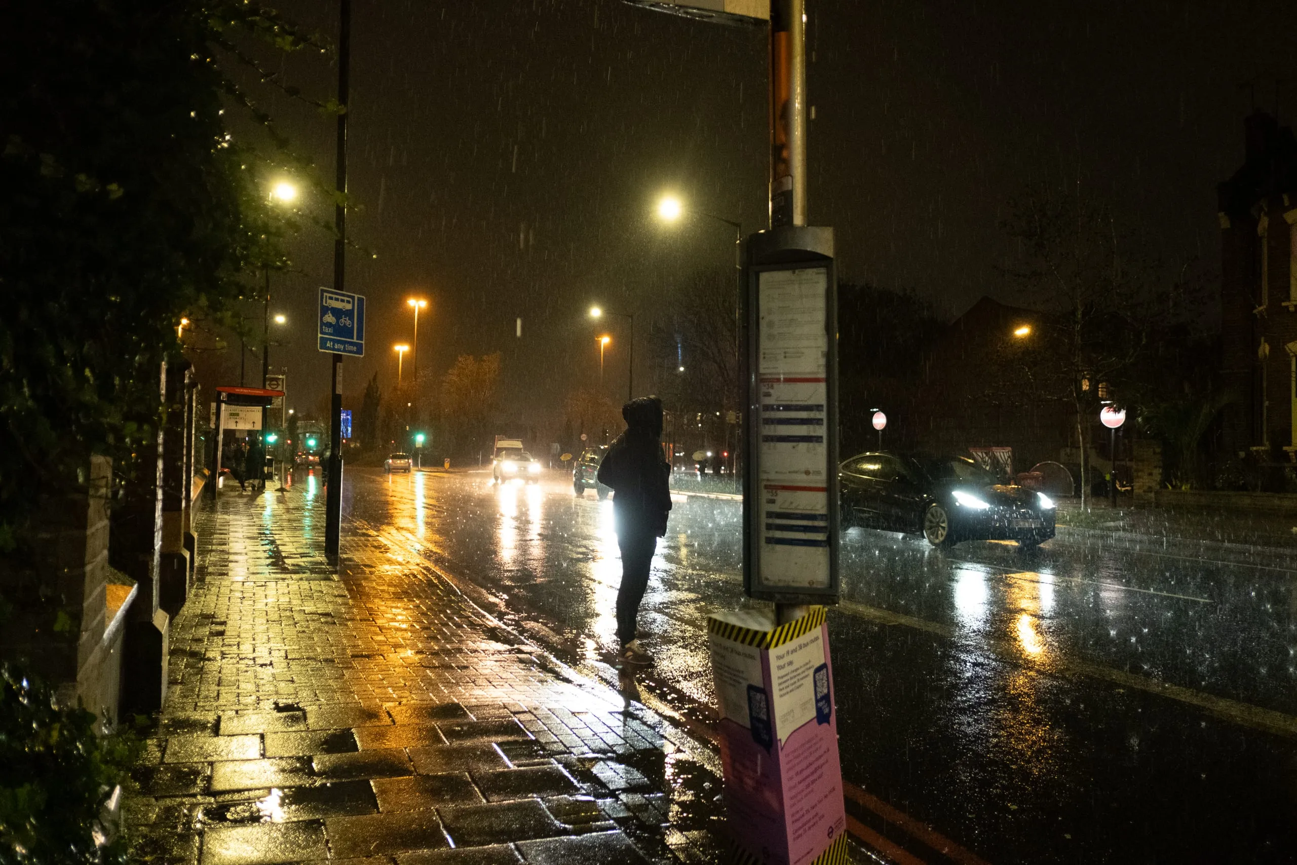 A figure crosses a wet road past a bus stop in the rain at night