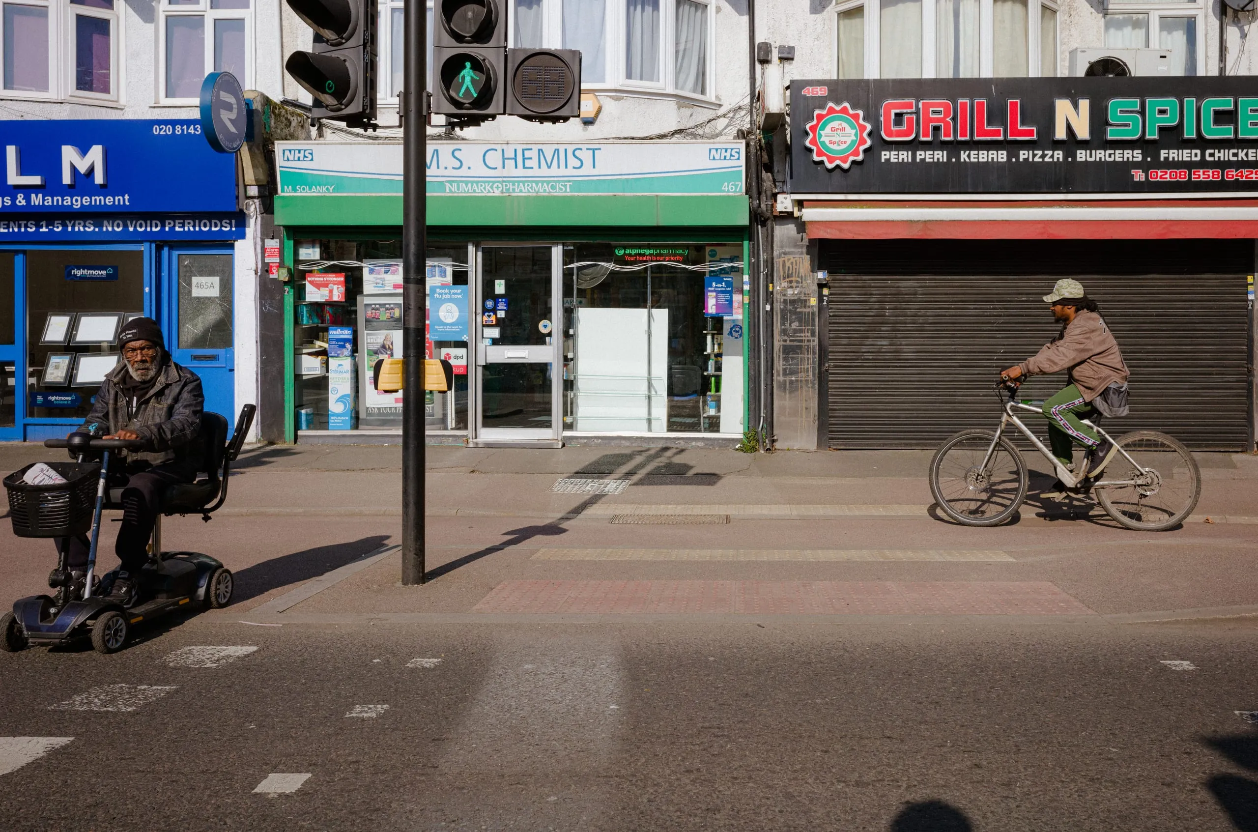 A man on a mobility scooter waits at a crossing as a cyclist passes a row of shops