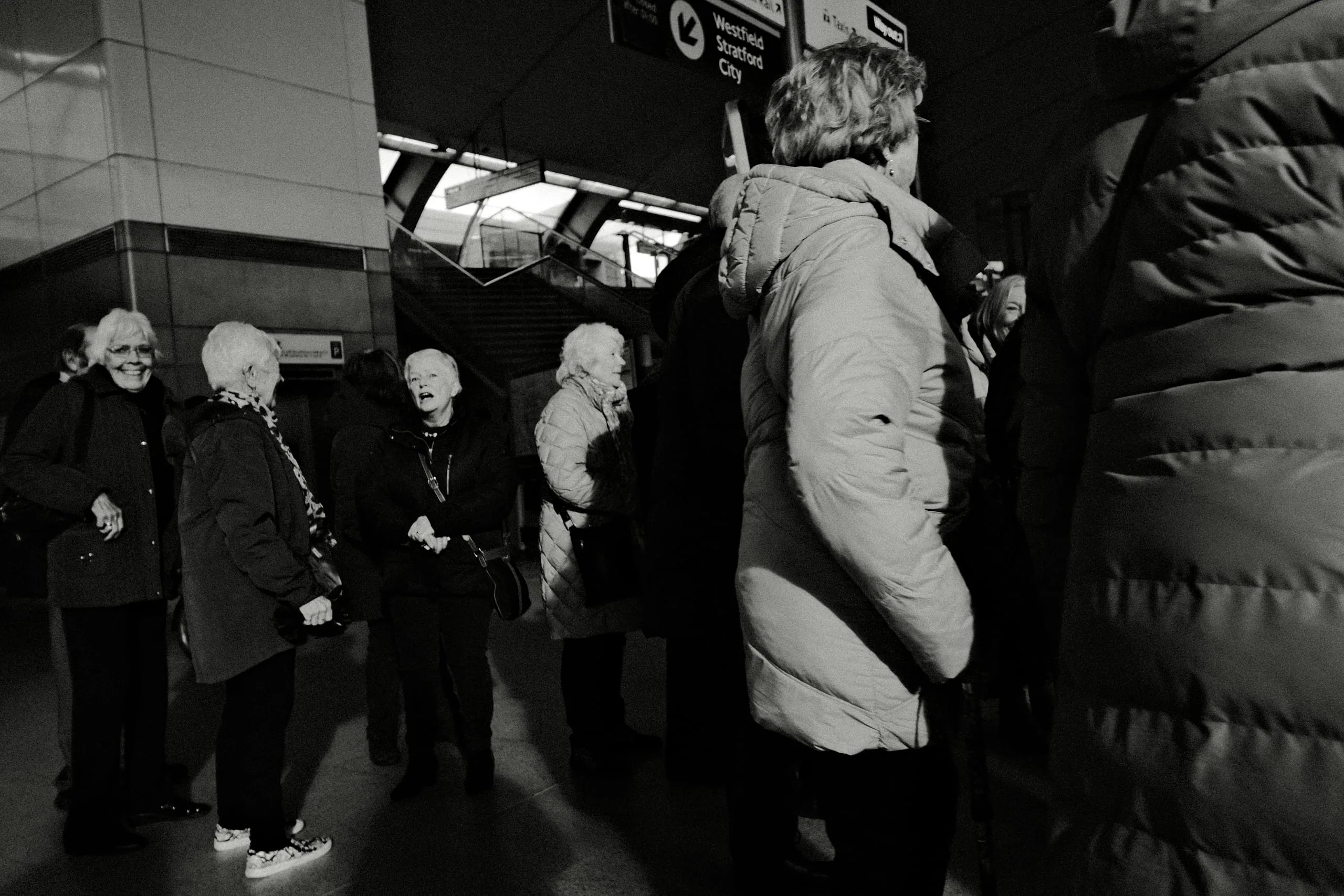 Older women talk on a station platform beneath a Westfield Stratford City sign, Stratford