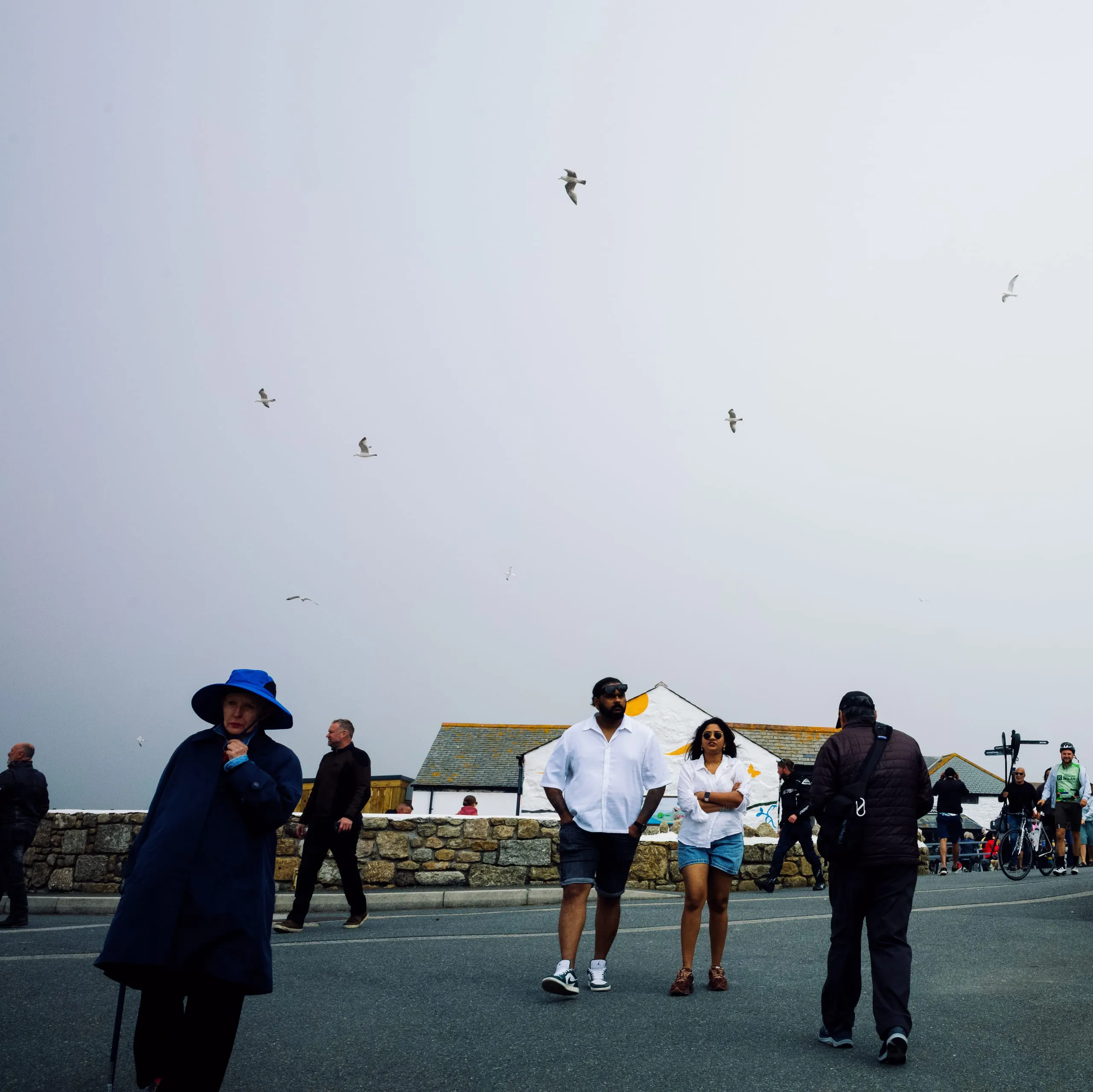 A woman in a blue hat walks towards the frame while seagulls overhead at Land's End