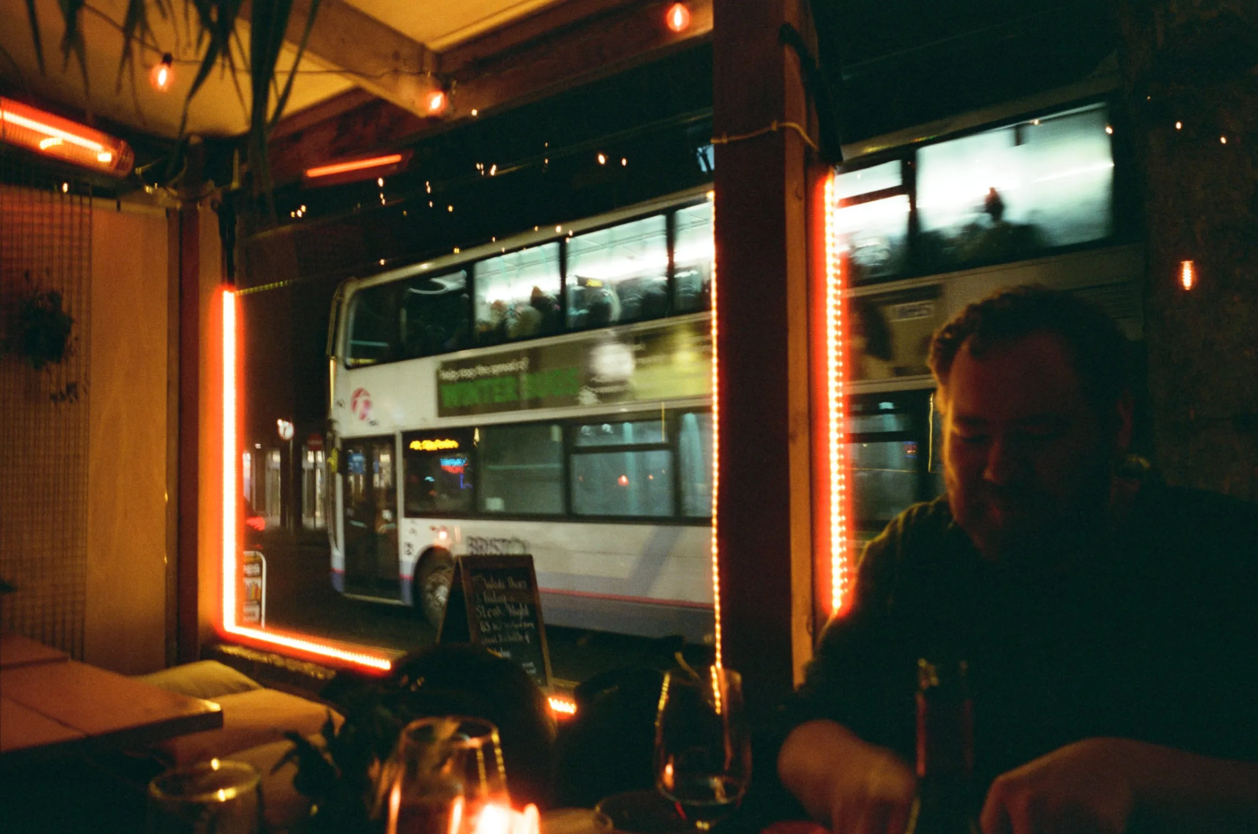A red double-decker bus passes the window of a bar at night