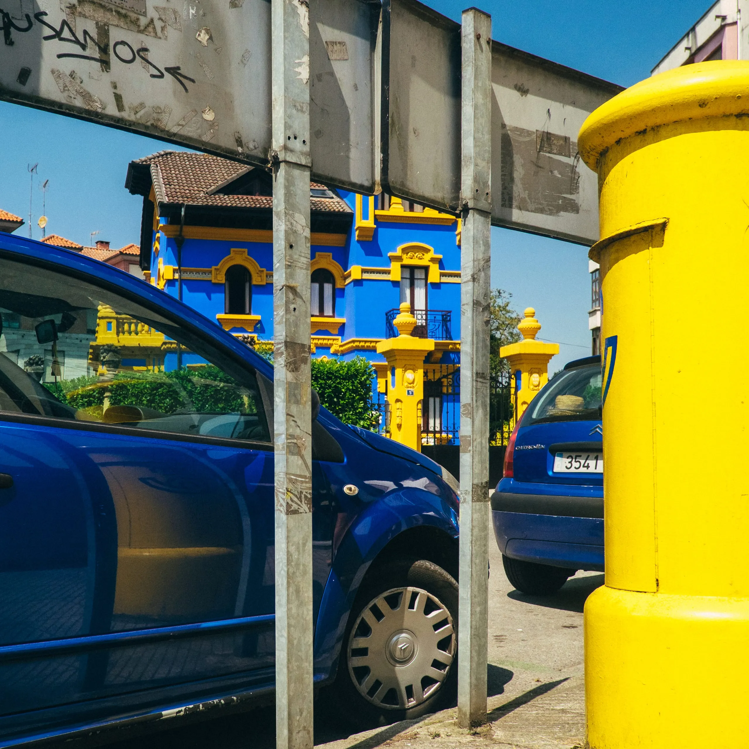 A blue tiled house seen past parked cars and a yellow Correos post box