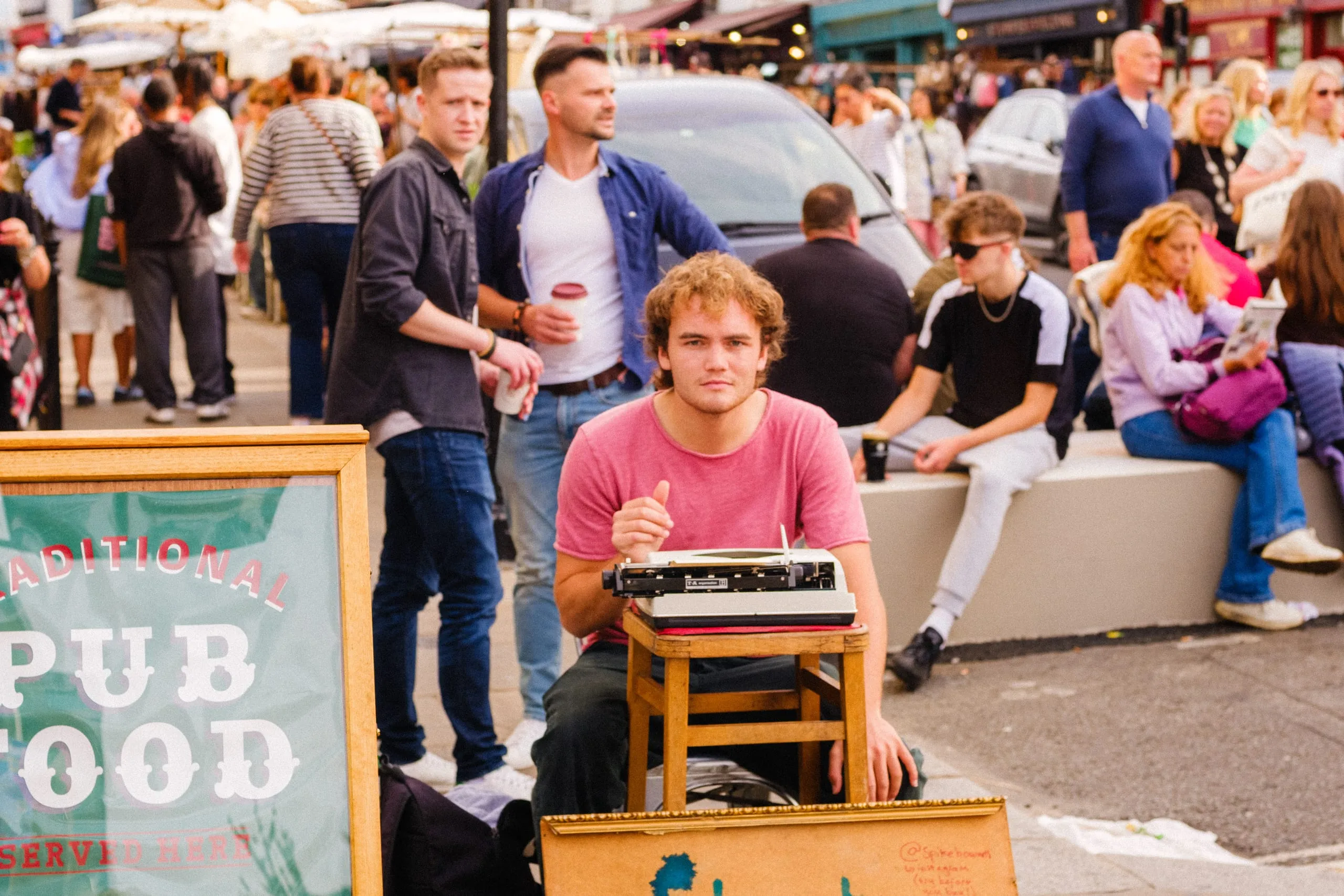 A young man sits at a typewriter on a street busy with people, Portobello Road