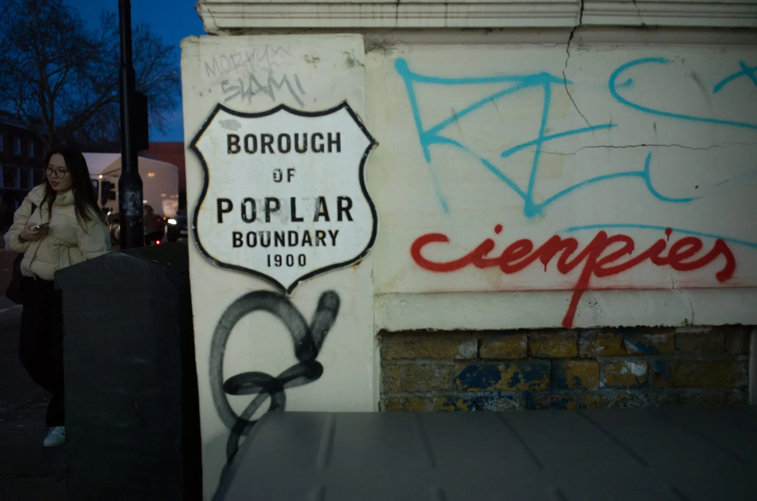 A Borough of Poplar boundary marker from 1900 beside graffiti, with a woman on her phone, Poplar