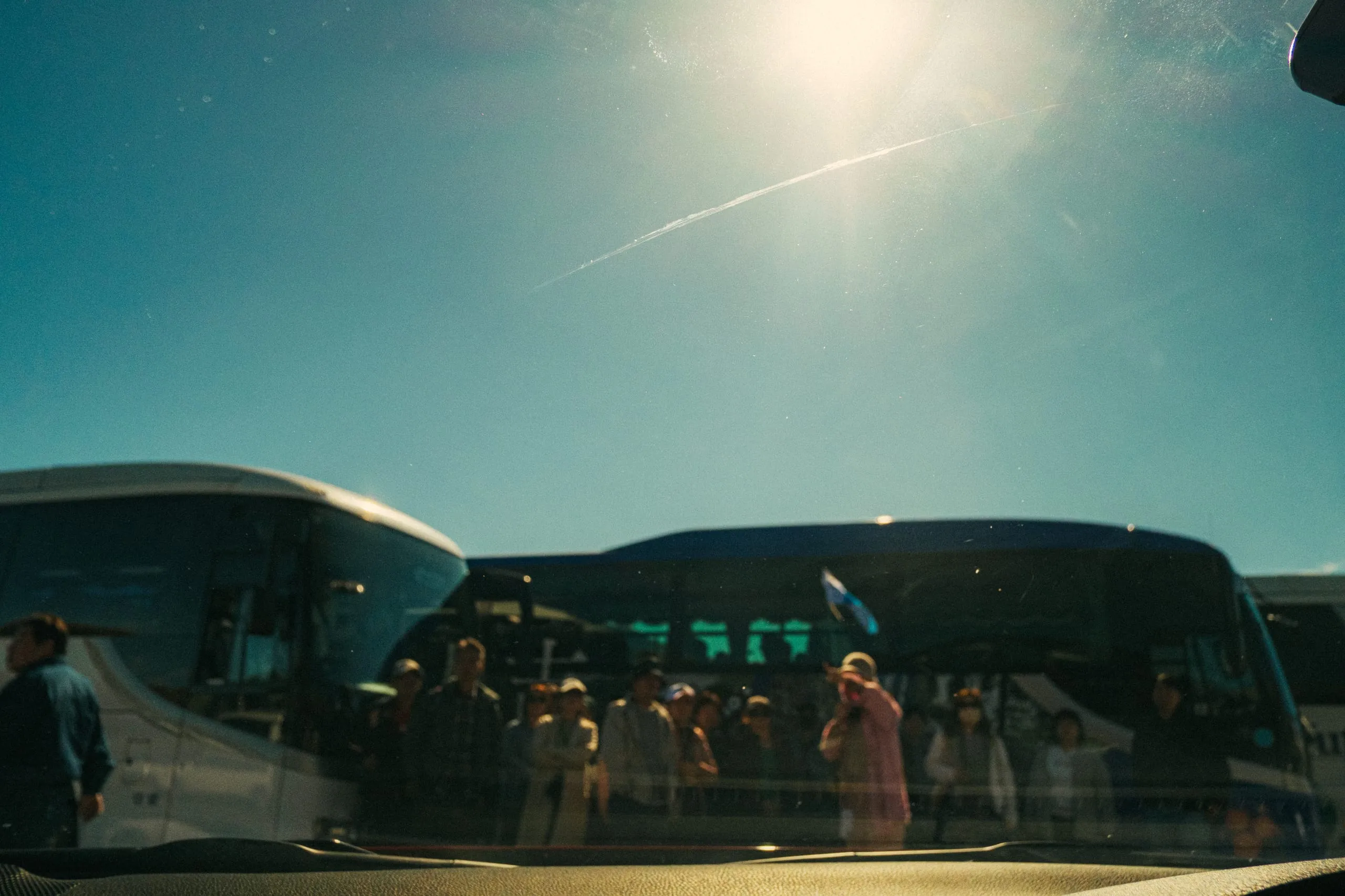 Tour groups gather beside parked coaches under a bright sky, seen through a windscreen