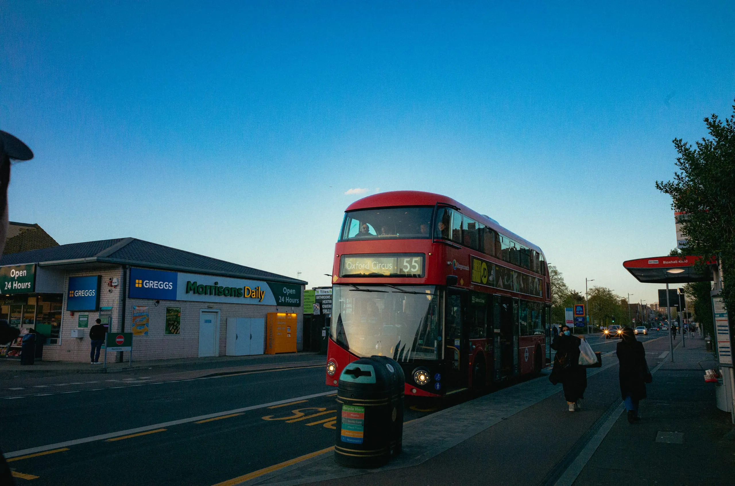 A route 55 double-decker bus waits at a stop beside a Greggs at dusk
