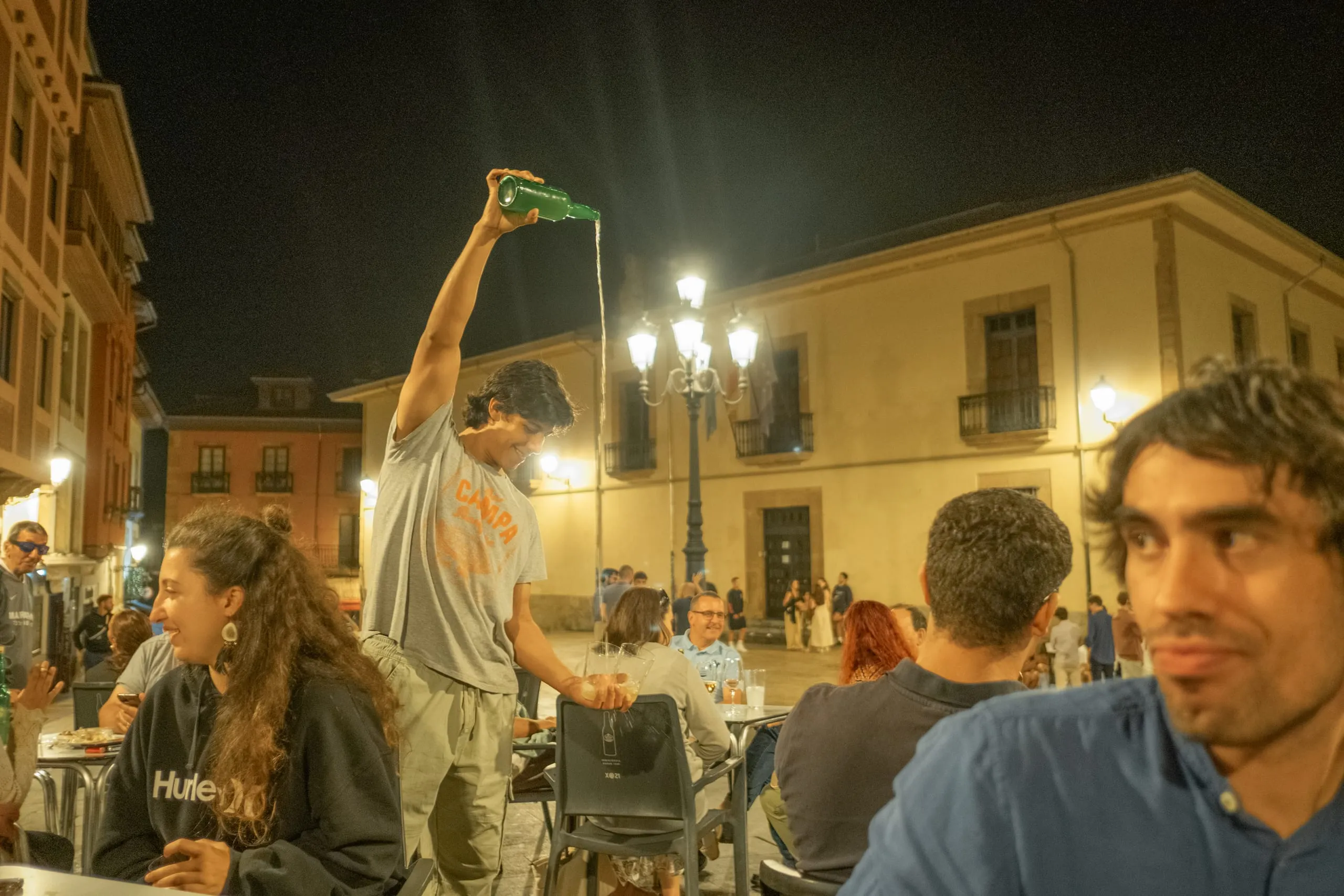 A man pours cider from a raised bottle at an outdoor table at night, Plaza del Campa