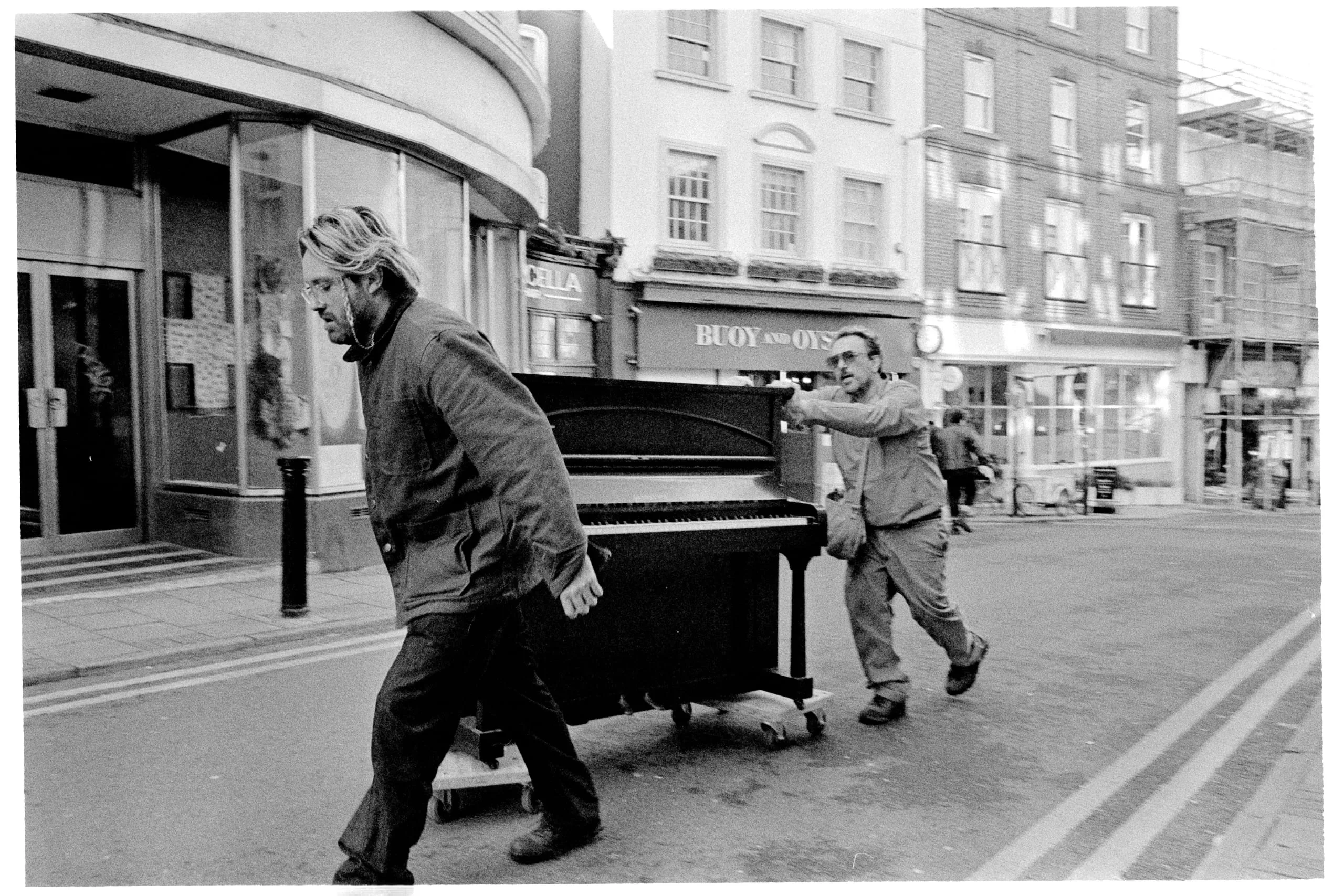 Two men move an upright piano across a street during Margate Off-Season art fair
