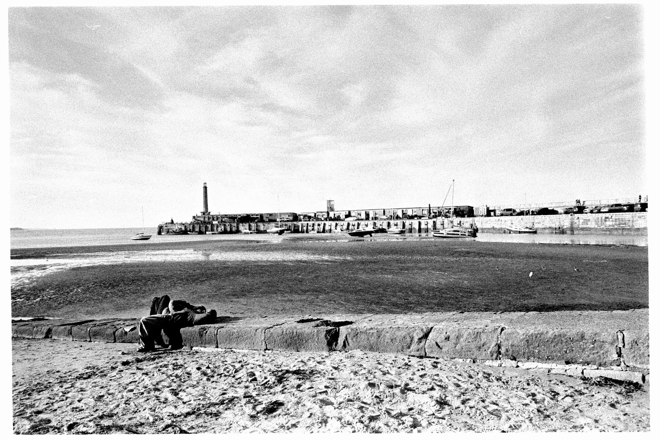 People sit on a sea wall above the beach, with the harbour and lighthouse behind, Margate