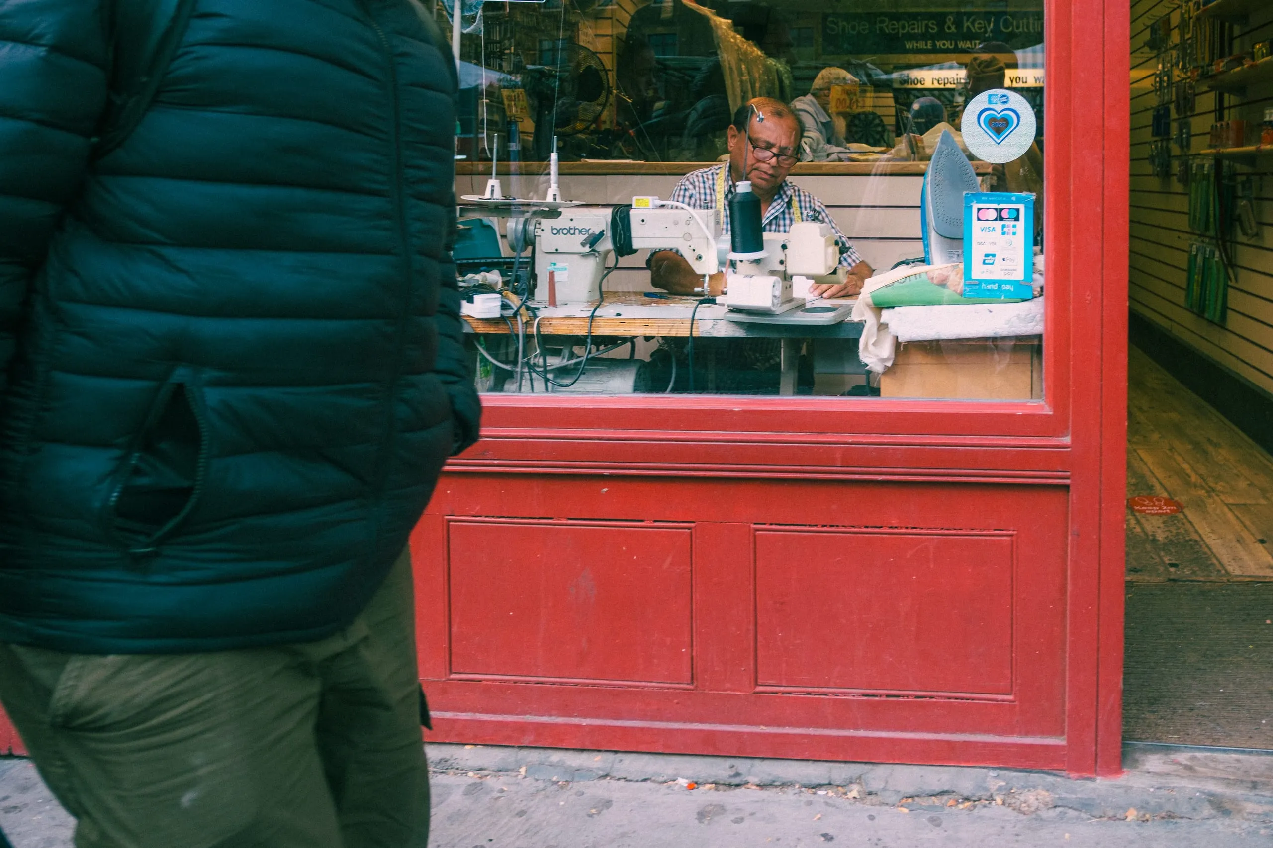 A man works at a sewing machine in the window of a shoe repair and key cutting shop, Whitechapel