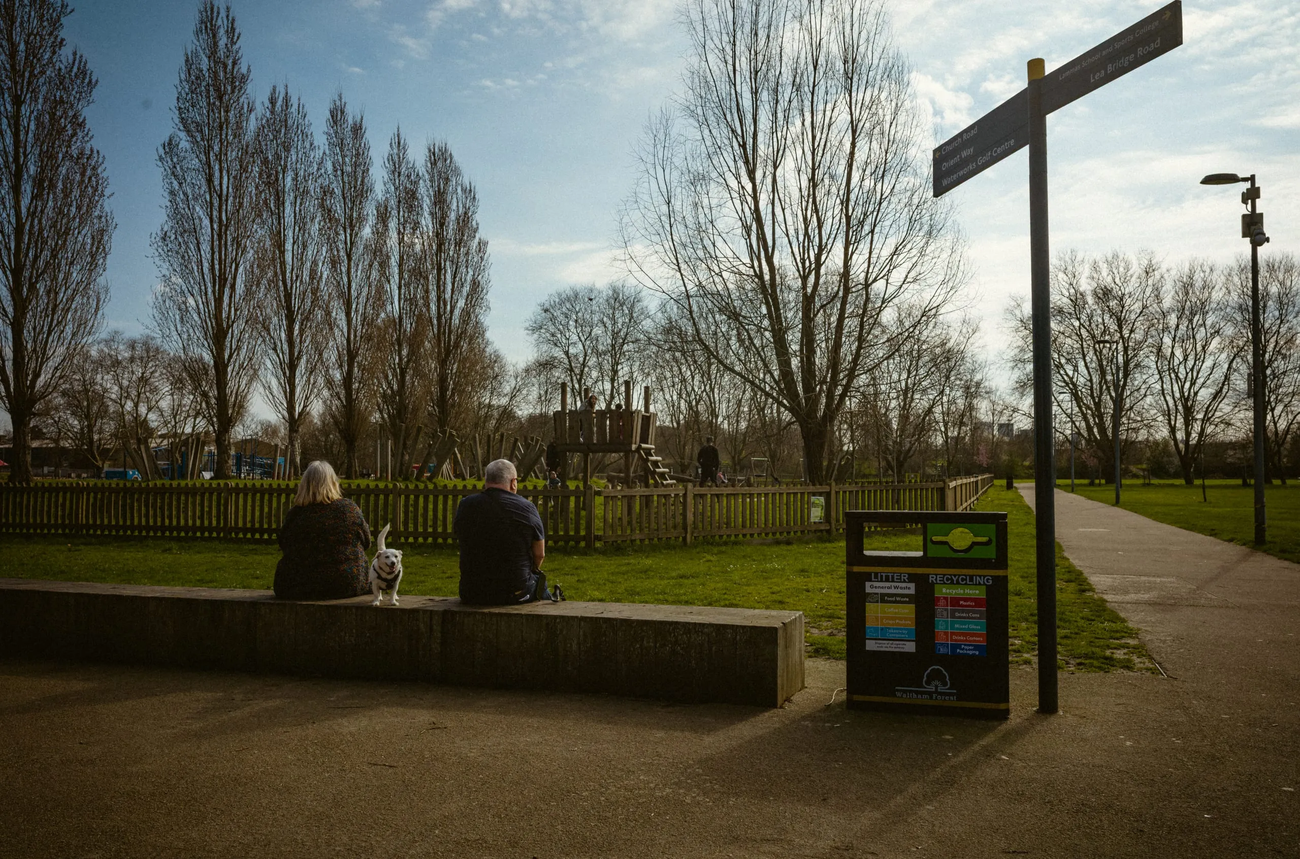 A couple sit with a dog on a wall at the edge of a park