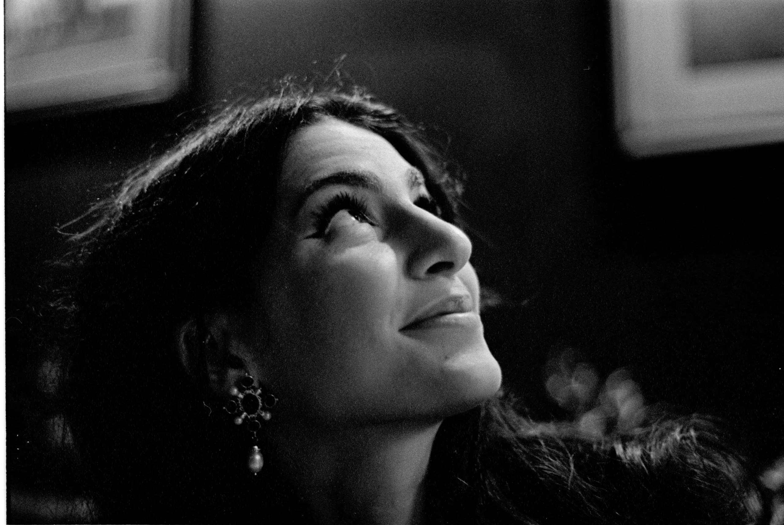 Caterina looks upward in a dimly lit room hung with framed pictures