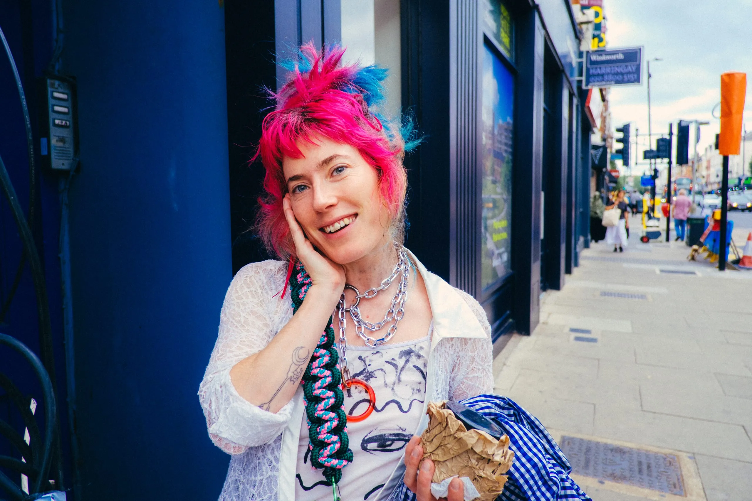 A woman with pink and blue hair leans against a shopfront, Harringay